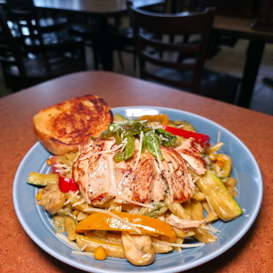 Grilled chicken and vegetables with peppers, zucchini, pasta, and cheese served on a blue plate in a restaurant.