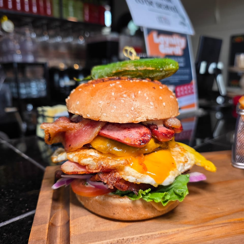 Towering bacon cheeseburger with a fried egg, sausage, vegetables, and pickles on a wooden serving board at a bar.