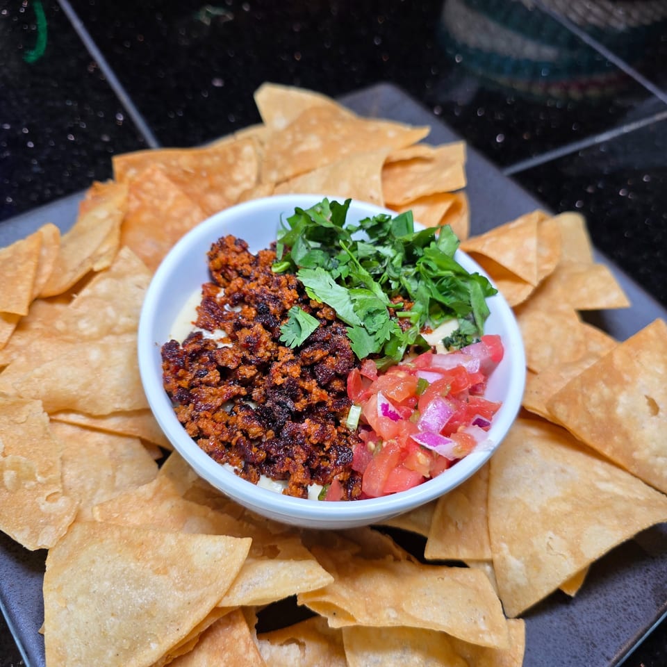 Bowl of creamy dip topped with spiced meat, herbs, and diced tomatoes, surrounded by crispy tortilla chips.