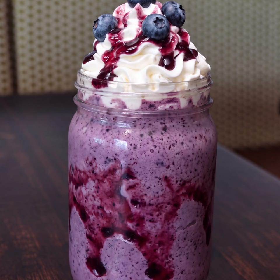 Blueberry smoothie topped with whipped cream, berry sauce, and fresh blueberries in a glass jar on a wooden table.