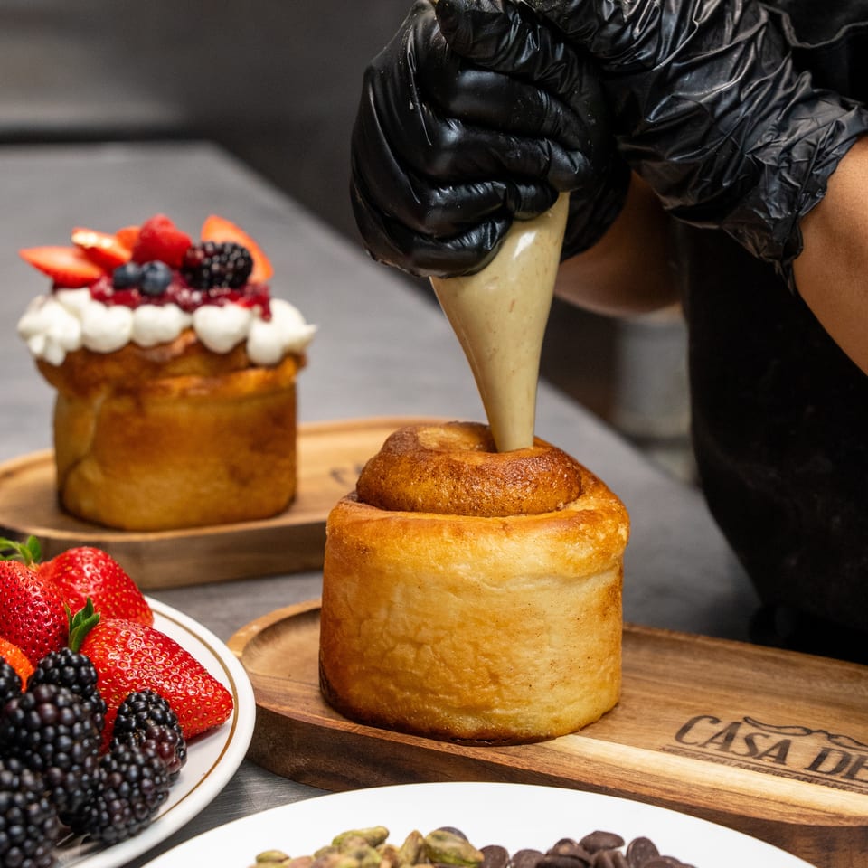 Gloved baker pipes cream into a pastry surrounded by fruit-topped desserts, berries, pistachios, and chocolate pieces.