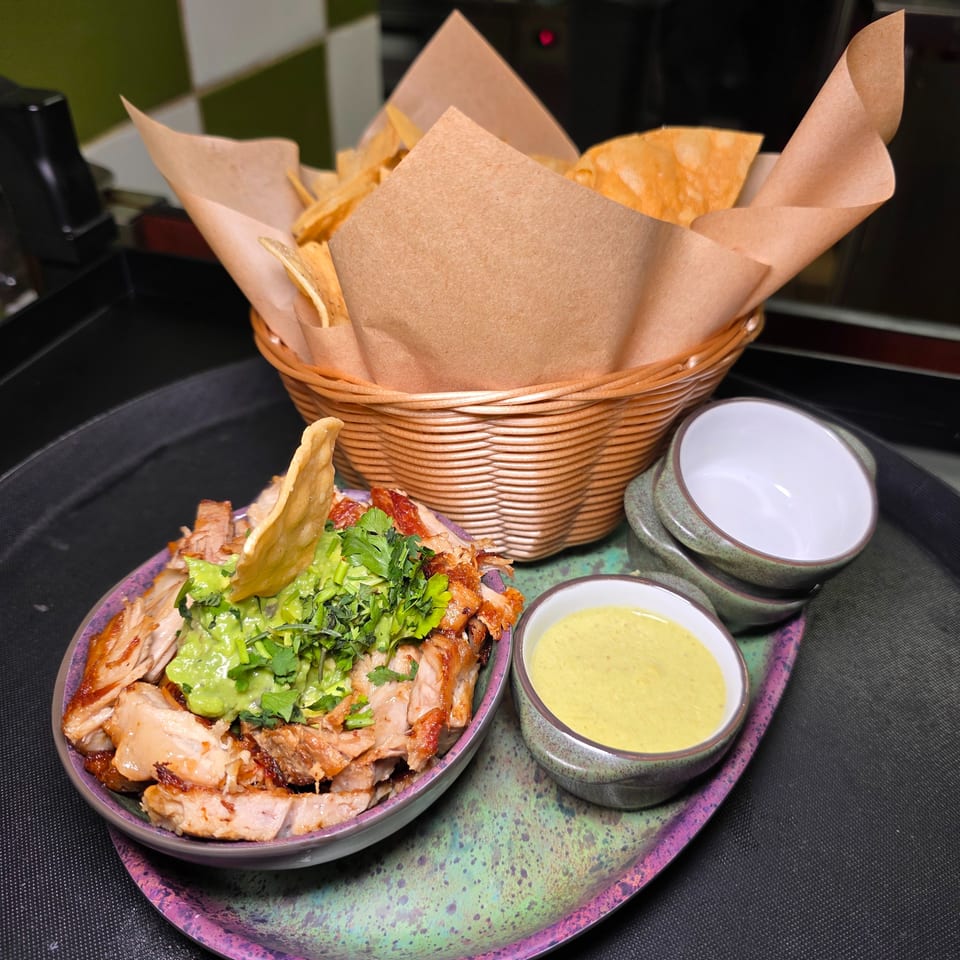 Basket of tortilla chips beside roasted meat topped with guacamole and herbs, served with dipping sauces on a colorful plate.