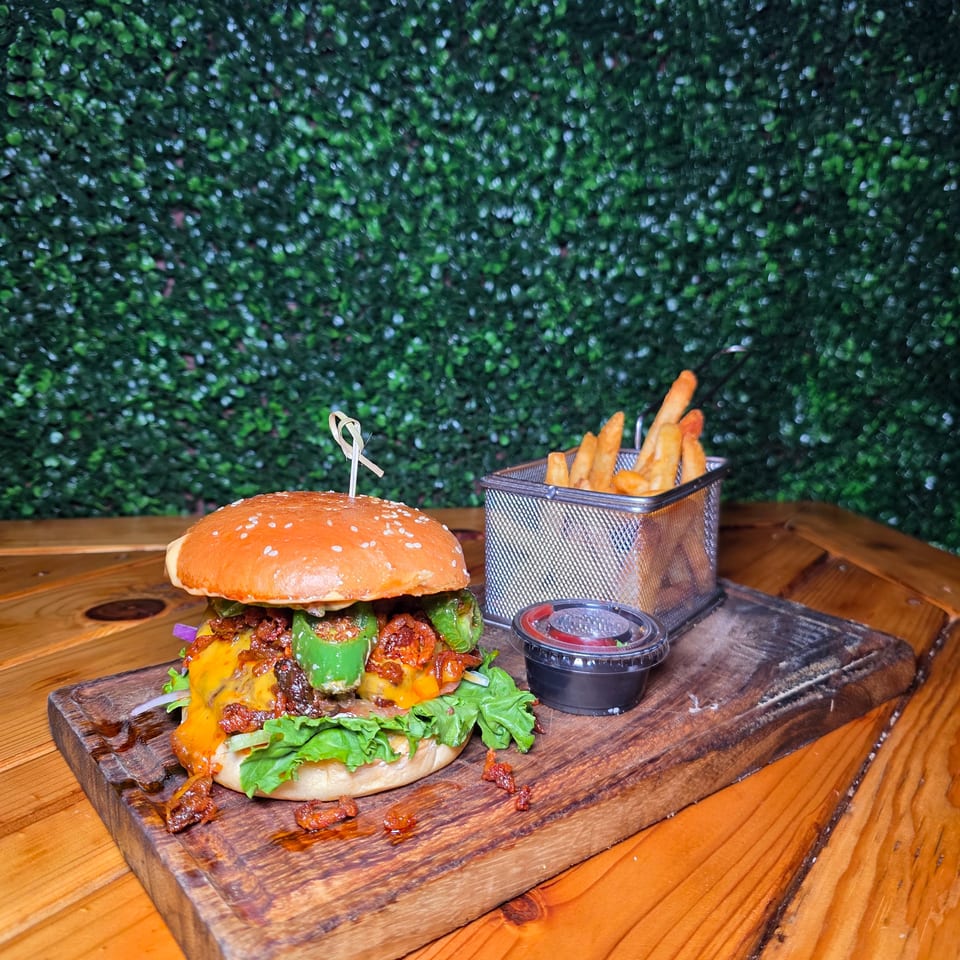 Loaded cheeseburger with lettuce, bacon, peppers, and sauce served on a wooden board with fries and dipping sauce.