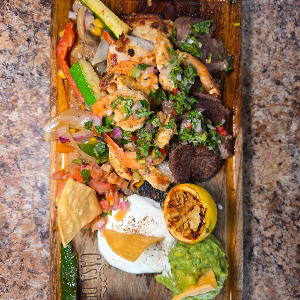 Wooden platter of grilled shrimp, chicken, steak, vegetables, salsa, guacamole, tortilla chips, and grilled citrus.
