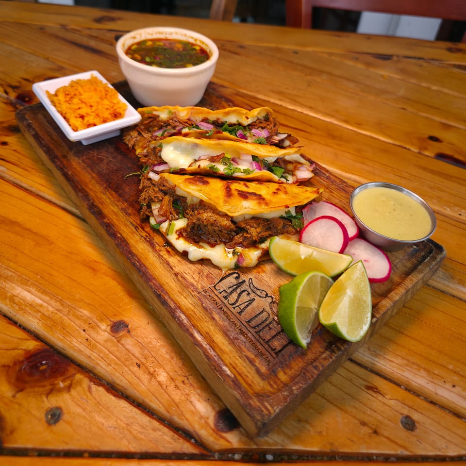 Three cheese-topped shredded-beef tacos with cilantro and onions served on a wooden board with rice, beans, sauces, radishes, and lime wedges.