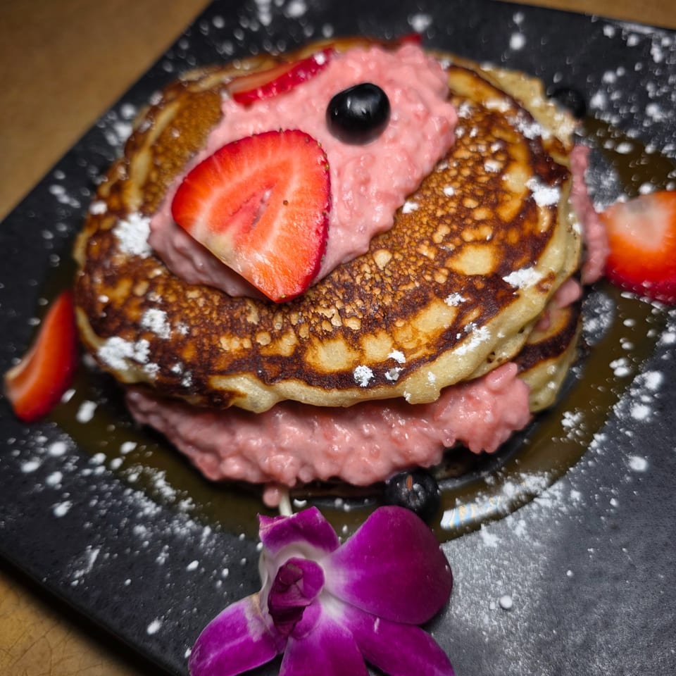 Stack of browned pancakes topped with pink cream, strawberries, blueberries, powdered sugar, and a purple orchid on a black plate.