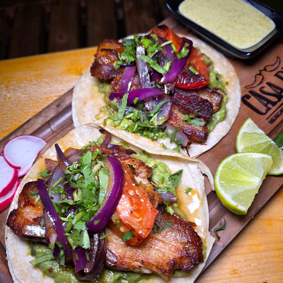 Two tacos topped with grilled meat, roasted onions, tomatoes, cilantro, and guacamole, served with lime wedges and creamy sauce.