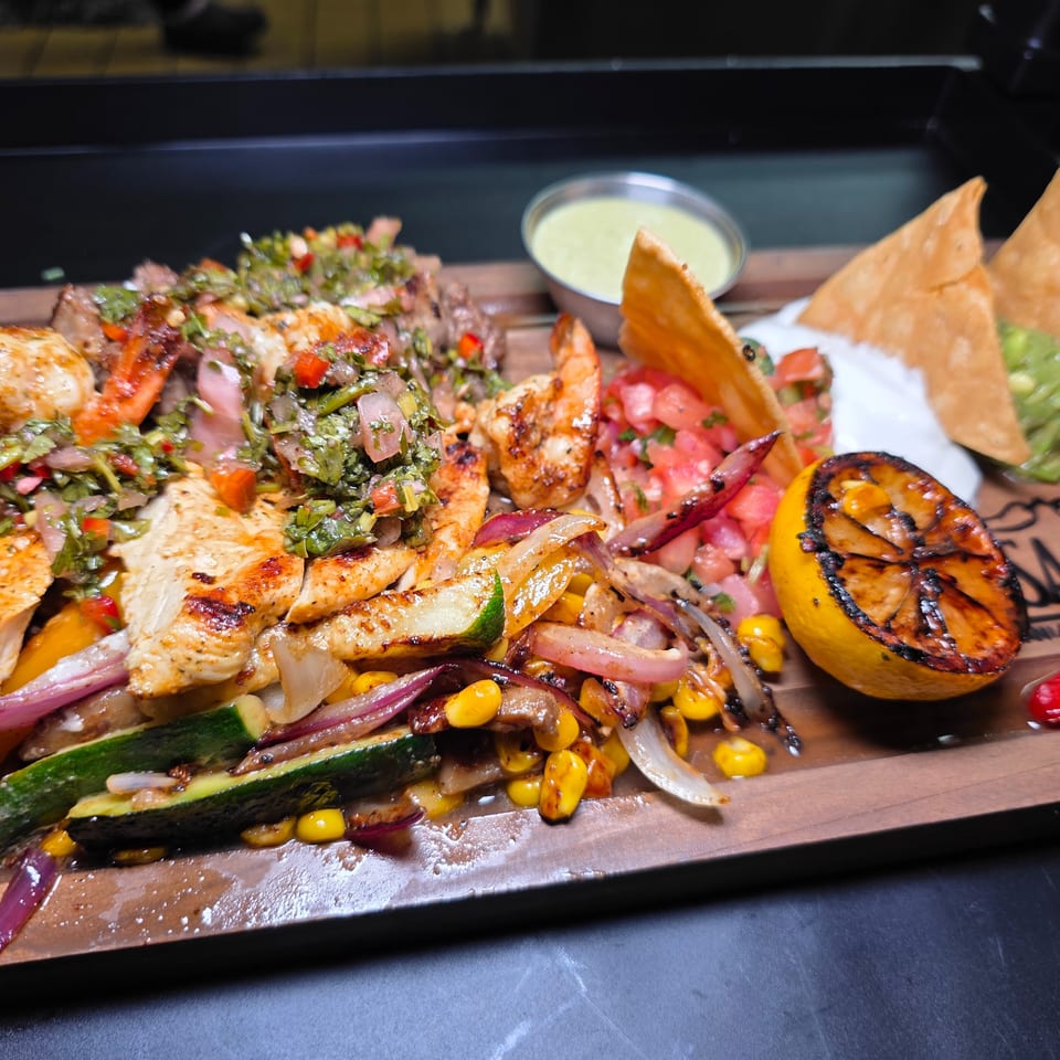 Grilled chicken, shrimp, vegetables, salsa, tortilla chips, guacamole, and dipping sauce arranged on a wooden serving board.