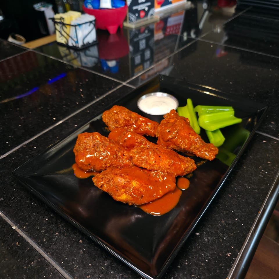 Buffalo chicken wings coated in orange sauce with celery sticks and ranch dip on a black plate at a restaurant counter.