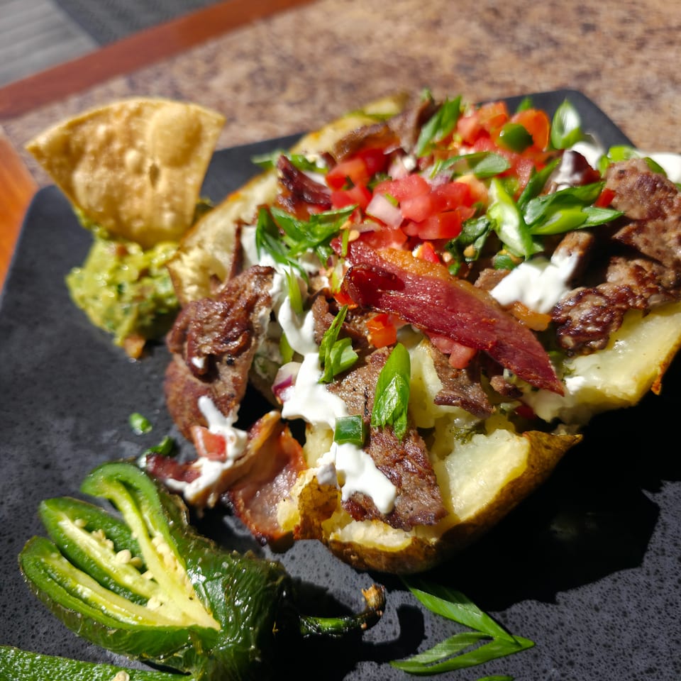 Loaded baked potato topped with grilled meat, bacon, tomatoes, herbs, jalapeños, and creamy sauce on a black plate.